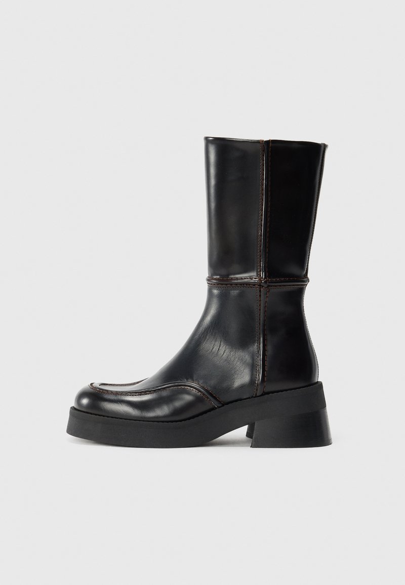 Bota de tornozelo em couro preto com salto bloco, apresentando costura em contraste e um design de bico curvo. Textura suave, silhueta elegante.