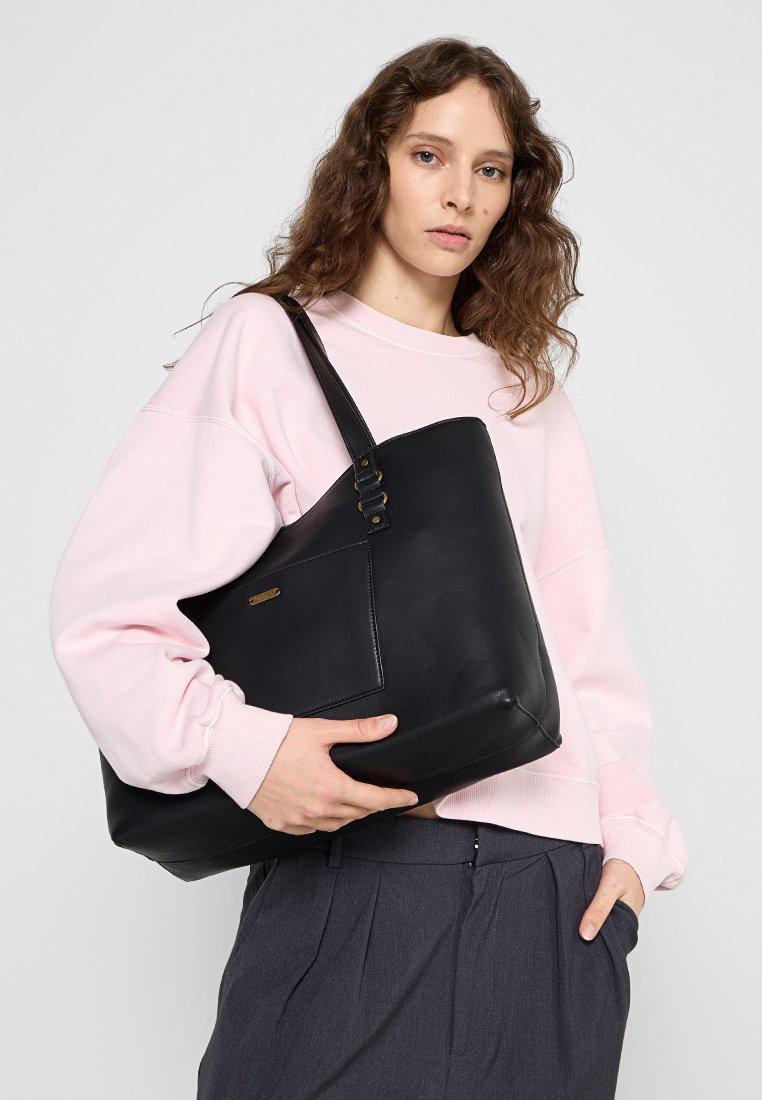 Bolso bandolera de cuero negro con una textura suave, un bolsillo delantero y una correa larga ajustable. Llevarlo una modelo con un sudadera de color rosa claro.
