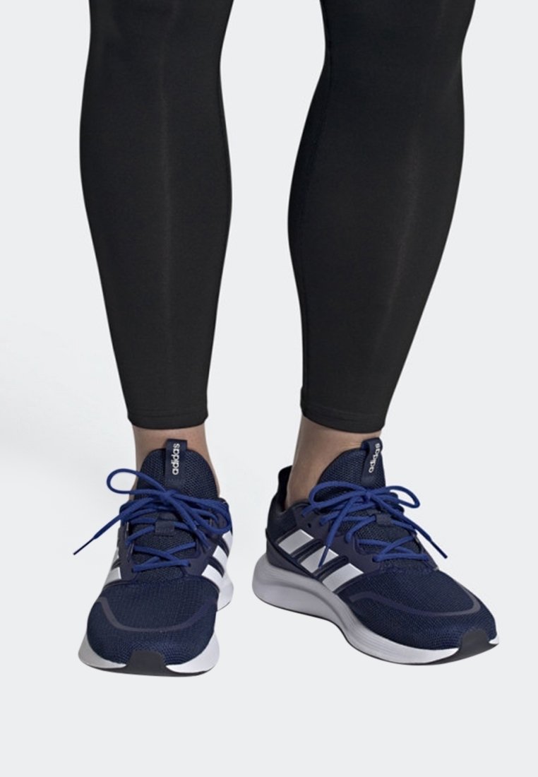 Personne portant un legging noir et des baskets Adidas bleu marine avec des bandes blanches et des lacets bleus, pieds tournés vers l'avant sur fond blanc.