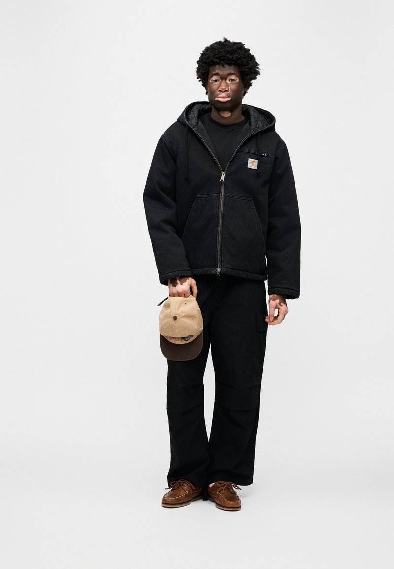 Sweat à capuche noir zippé avec un logo sur le devant, porté avec un pantalon cargo noir. Tenant une casquette de baseball beige et marron, portant des chaussures bateau marron.