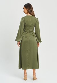 Olivno zelena obleka z dolgimi naborki, prilegajočim bodijem in tekočo krilom. Ima zadrgo na hrbtni strani in gladko, mat teksturo.