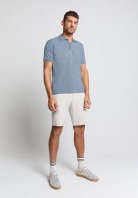 Lichtblauw poloshirt met korte mouwen, gecombineerd met beige shorts. Model draagt grijze sneakers met witte accenten en gestreepte sokken.