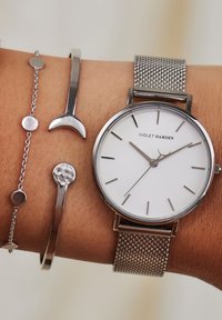 Zilveren mesh horloge met een witte wijzerplaat en minimalistische zilveren uurmarkeringen, vergezeld door twee zilveren armbanden met verschillende ontwerpen.