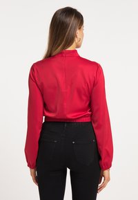 Bluză din satin roșu cu guler înalt, mâneci lungi bufante și tiv elastic. Închidere cu fermoar la spate. Asortată cu blugi negri cu talie înaltă.