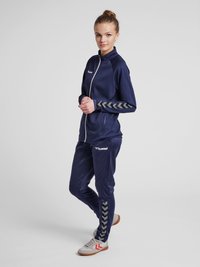 Marineblauw sporttracksuit met een ritsjack en bijpassende broek. Bevat grijze chevron-accenten op de mouwen en de broek. Gemaakt van een zachte stof.