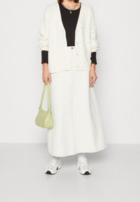 Vit stickad cardigan med strukturerat mönster över en svart topp, tillsammans med en lång vit kjol, en grön handväska och vita sneakers.