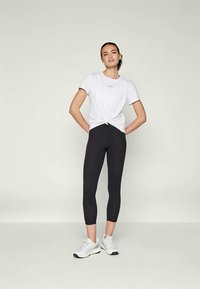 Balta T-kreklu ar mezglošanas detaļu viduklī, apvienots ar melniem leggings un pelēkiem sporta apaviem, uz neitrālas fona.