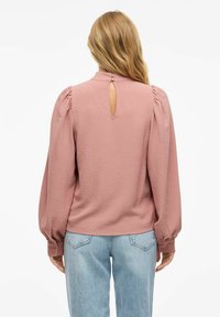 Lätt texturerad, dammig rosafärgad blus med puffärmar, ett nyckelhål bak, och knappade manschetter, ihop med ljusblå jeans.