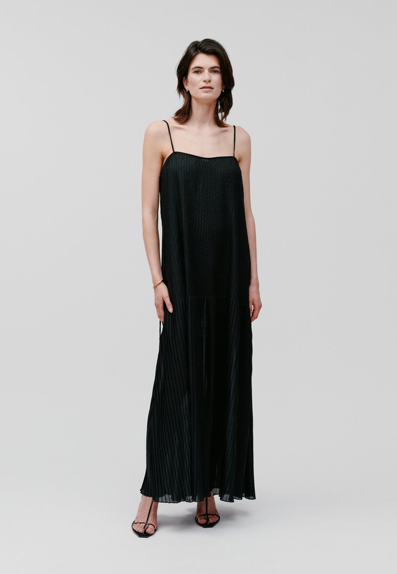 Black pleated maxi dress with thin spaghetti straps, featuring a textured bodice and flowing skirt, paired with black strappy heels.