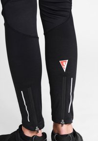 Schwarze Radfahrleggings mit reflektierenden Akzenten, Reißverschlüssen an den Knöcheln und einem Logo-Patch in Rot und Weiß an der Seite.