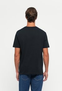 Mand med kort bølget hår iført en ensfarvet sort kortærmet t-shirt og blå jeans, stående vendt væk med en hvid baggrund.