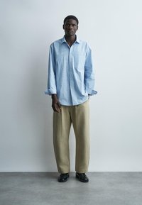 Férfi, aki túlméretezett, világoskék csíkos inget, bézs színű, bőszárú nadrágot és fekete cipőt visel, miközben egy sima, világos falnak támaszkodik.