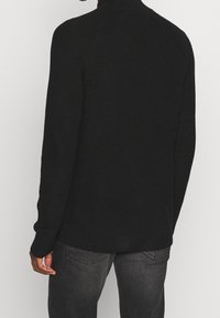 Svart stickad tröja med hög krage, med texturerad tyg och långa ärmar, bärs tillsammans med mörkgrå jeans.