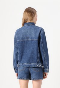 Denim jacket in dark blue with a collar, long sleeves, and silver snap buttons. Features visible stitching and a logo on the back.