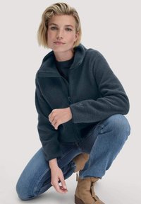 Giacca in pile grigio scuro con colletto alto e chiusura a zip, abbinata a jeans blu e stivali alla caviglia color sabbia. Tessuto morbido e vestibilità comoda.