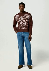 Vyras, vilkintis rudą megztinį su gamtos peizažu, mėlynus džinsus ir rudus batus, stovintis su viena ranka kišenėje prieš paprastą foną.