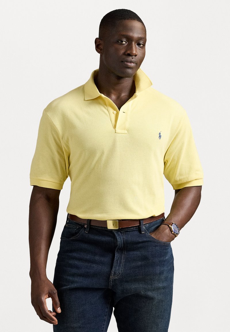 Camisa polo amarela com gola e mangas curtas, combinada com jeans escuros e um cinto castanho. Apresenta um pequeno logótipo.