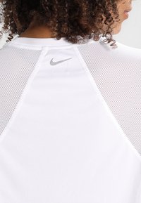 Mujer con cabello rizado que lleva una camiseta blanca de Nike con paneles de malla y un logotipo gris de Nike en la parte superior de la espalda.