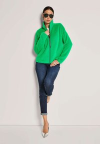 Frau in einer leuchtend grünen, flauschigen Jacke, dunkler hochgerollter Jeans, weißen spitzen High Heels, schwarzen Sonnenbrillen und silbernen Ohrringen, die vor einer schlichten Wand posiert.