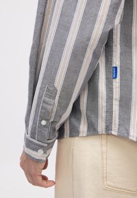 Cămașă pe dungi gri și bej, cu mâneci lungi și mansete cu nasturi, având un eticheta logo albastră și o croială relaxată. Materialul pare texturat și ușor.