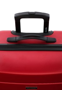 Maleta rígida roja con superficie texturizada, cremallera negra y asa. Cuenta con un asa retráctil montada en la parte superior y una placa con el logo de la marca.