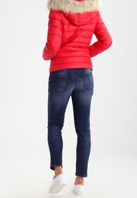 Czerwona pikowana kurtka z futrzanym kapturem, noszona na ciemnoniebieskie dopasowane dżinsy i białe trampki, widok z tyłu.