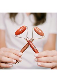 Two red facial rollers with shiny, glittery handles and gold metal frames, held by hands with neutral nail polish.