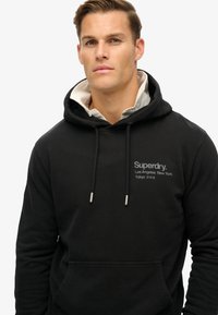 Sudadera negra de tela suave, con capucha ajustable, bolsillo frontal y logo en texto gris. Capa con una camiseta de color claro debajo.