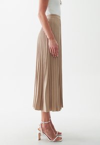 Gonna midi plissettata beige con una texture a coste, abbinata a sandali alti bianchi con cinturini. Vista di lato che mostra il design e il movimento del tessuto.
