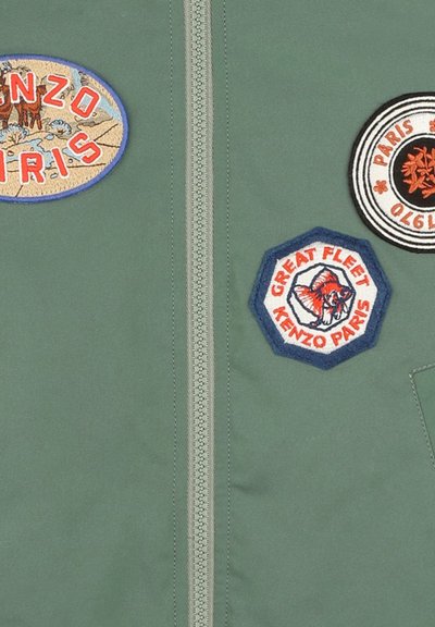Veste en tissu vert avec une fermeture éclair centrale. Présente trois patchs brodés : oval 'KENZO PARIS', rond noir et blanc, et hexagonal 'GREAT FLEET'.