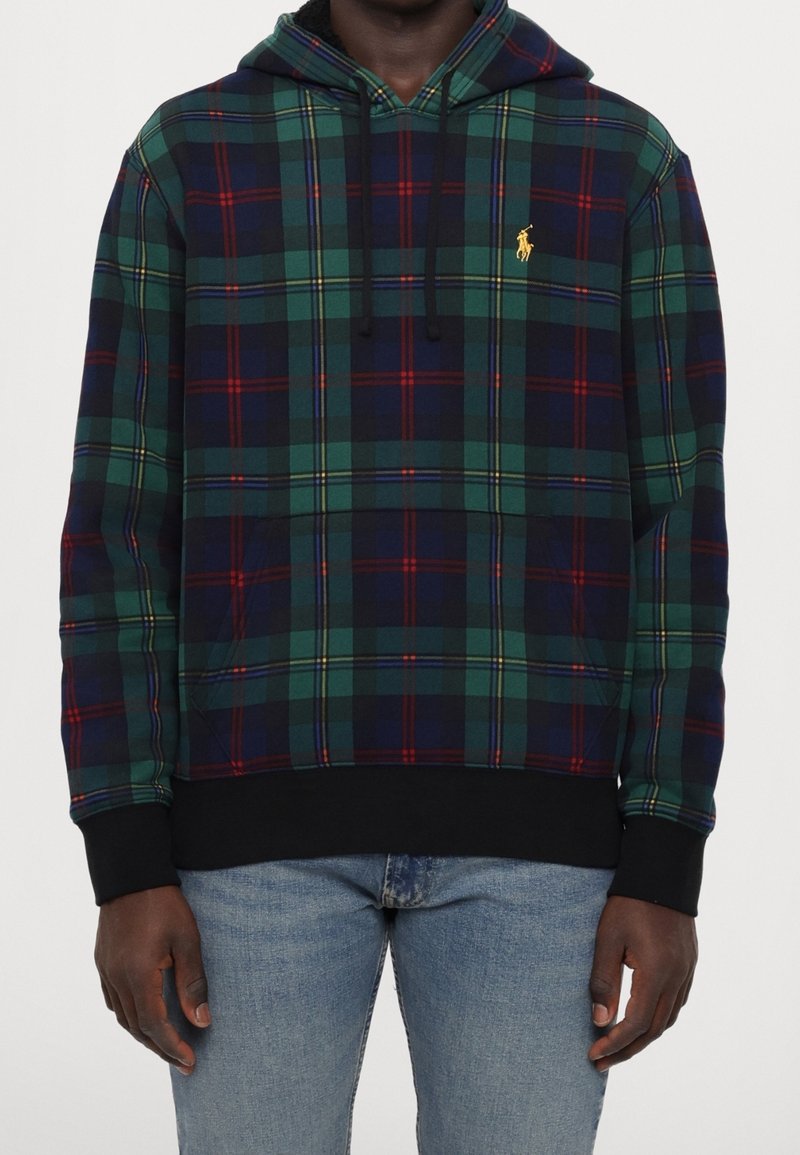 Personne portant un sweat à capuche à carreaux verts, bleu marine et rouge avec des poignets et un ourlet noirs, associé à un jean bleu clair, debout devant un fond uni.