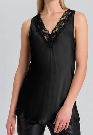 Kobieta w czarnej atłasowej bluzce bez rękawów z dekoltem w kształcie litery V wykończonym czarną koronką, w połączeniu z czarnymi skórzanymi spodniami.