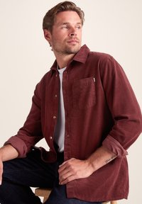 Burgundy corduroy shirt with long sleeves, front pocket, and button closure. Worn over a white shirt, paired with dark jeans.
