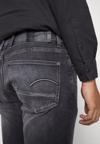 Jeans de mezclilla negra con un acabado desgastado, cinco bolsillos, costuras de contraste en el bolsillo trasero y un parche de cuero con marca en la cinturilla.