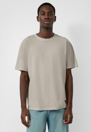 Jonge man met een donkere huid draagt een beige oversized T-shirt met korte mouwen en lichtblauwe spijkerbroek, staand tegen een effen lichte achtergrond.