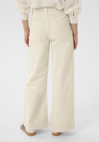 Pantalon crème à jambes larges avec une texture lisse, doté de deux poches arrière et d'une taille haute, associé à des chaussures de couleur claire.