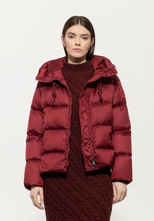Burgundy puffer jacket with a hood, zip front, and drawstring accents. Features a quilted design and smooth texture. Worn over a knitted sweater.