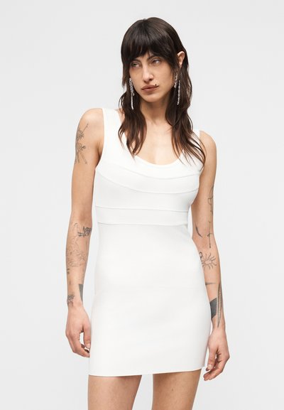 Νεαρό άτομο με μακριά σκούρα μαλλιά και τατουάζ και στα δύο μπράτσα, που φοράει στενή λευκή αμάνικη φορεσιά, σε απλό φόντο.