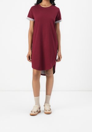 Kobieta ubrana w bordową sukienkę z krótkim rękawem z szarymi podwiniętymi mankietami, beżowe skarpetki i beżowe trampki w kwiatowy wzór na białym tle.