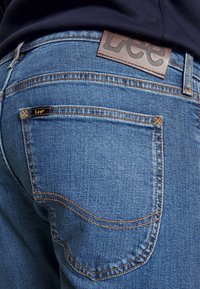 Jeans i denim med en medelblå tvätt, med gyllene sömmar, detaljer på bakfickorna och en brun läderlogotyp i närheten av midjebandet.