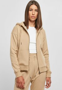Beige zip-up hettegenser laget av mykt materiale, med dobbel foret hette, frontlommer og snor detaljer. Kombinert med en hvit topp.
