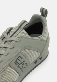 Scarpa sportiva grigia con una tomaia in mesh testurizzato, dettagli in gomma liscia, logo laterale e suola di un colore scuro a contrasto.