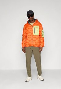 Orange puffer jacket with green and gray accents, featuring a front pocket and hood. Olive pants and white sneakers complete the look.