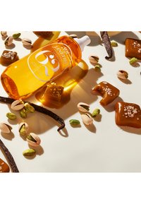 Bottiglia ambra del profumo Sol de Janeiro con etichetta bianca, circondata da pistacchi verdi e marroni, pezzi di caramello e bacche di vaniglia.