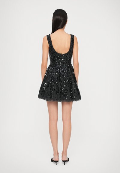 Žena s dlouhými tmavými vlasy je oblečena v bezramínkových černých mini šatech s flitry a černých lodičkách, stojí otočená zády na jednobarevném pozadí.