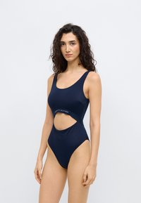 Ολόσωμο μαγιό σε ναυτικό μπλε χρώμα με ψηλή κοπή στο πόδι, λαιμόκοψη σε σχήμα scoop και σχεδιασμό με ανοίγματα, με ζώνη μέσης με επωνυμία. Λείο υλικό.