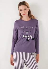 Ljubičasti longsleeve s grafikom medvjeda koji nosi prugasti šal; natpis "zima dolazi". Prugaste hlače u ljubičastoj i bijeloj boji. Mekani materijal.