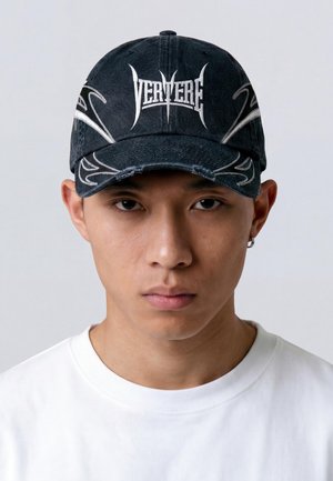 Joven con gorra negra con diseños tribales blancos y texto "VERITERE", camiseta blanca lisa, expresión neutral, fondo gris.