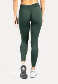 Leggings met hoge taille, nauwsluitend, in donker groene stof met een gladde textuur. Voorzien van contrasterende stiksels en gecombineerd met witte sportschoenen.