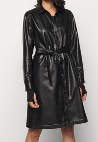 Černé kožené šaty košile s límečkem, předními kapsami a zavazováním v pase. Mají dlouhé rukávy s žebrovanými manžetami a knoflíky po celé délce.